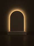 Volta lamp: a stone-toned arch on a plinth with a warm halo glowing on the wall behind it